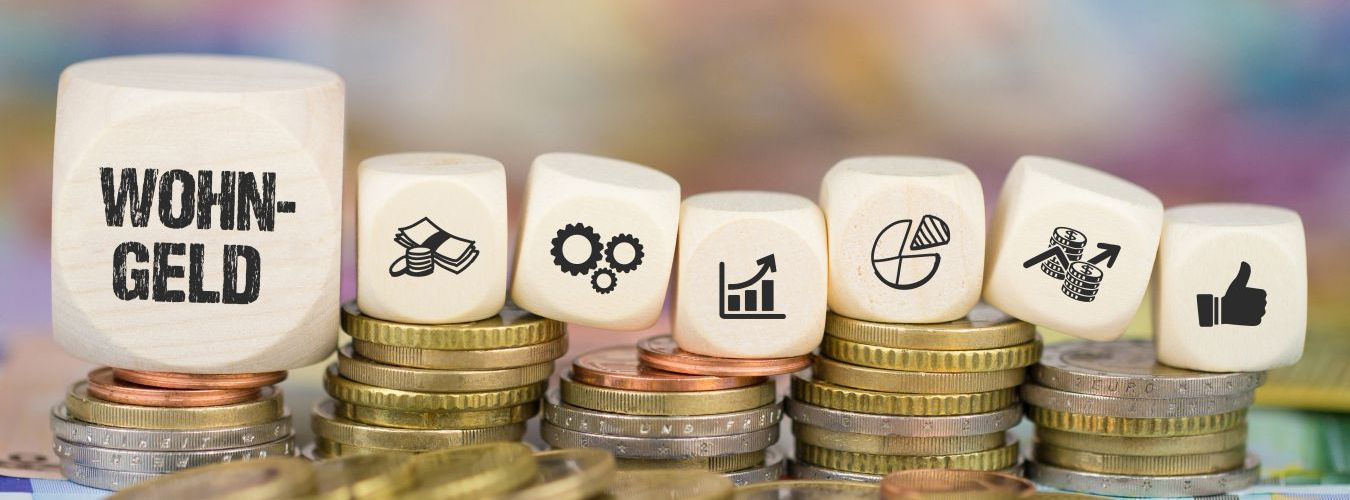 Holzwürfel mit der Aufschrift ‚Wohngeld‘ sowie Würfel mit Symbolen für Geld, Zahnräder, Wachstumsdiagramme und Daumen-hoch stehen auf gestapelten Euromünzen
