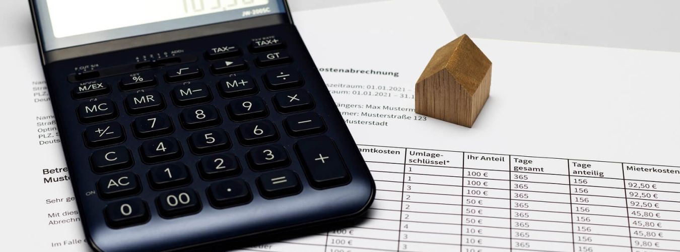 Taschenrechner mit Nebenkostenabrechnung und Modellhaus aus Holz auf einem Schreibtisch