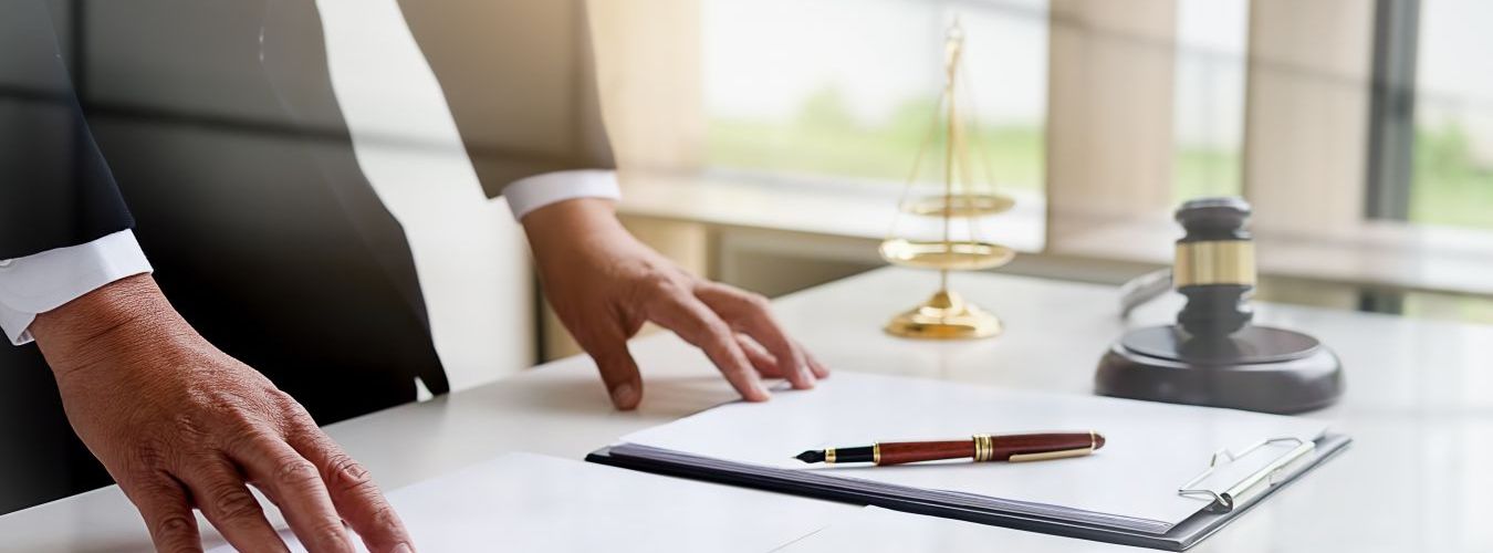 Mann am Schreibtisch mit Notizblock, im Hintergrund Waage der Gerechtigkeit und Richterhammer